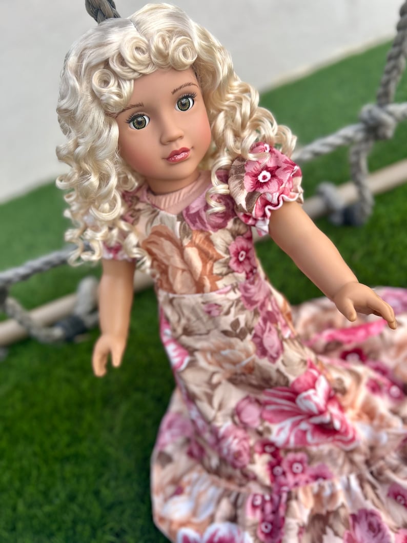 May include: A doll with blonde curly hair wearing a pink and brown floral dress. The doll has green eyes and is looking at the camera.