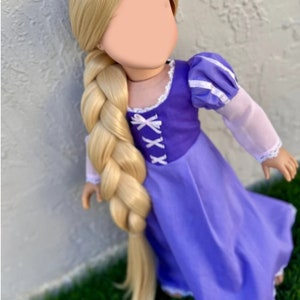 May include: A doll with long, blonde hair styled in a thick braid. The doll wears a purple dress with white accents and a bodice with criss-cross lacing. The sleeves are light pink with white trim. The doll is posed against a textured wall.