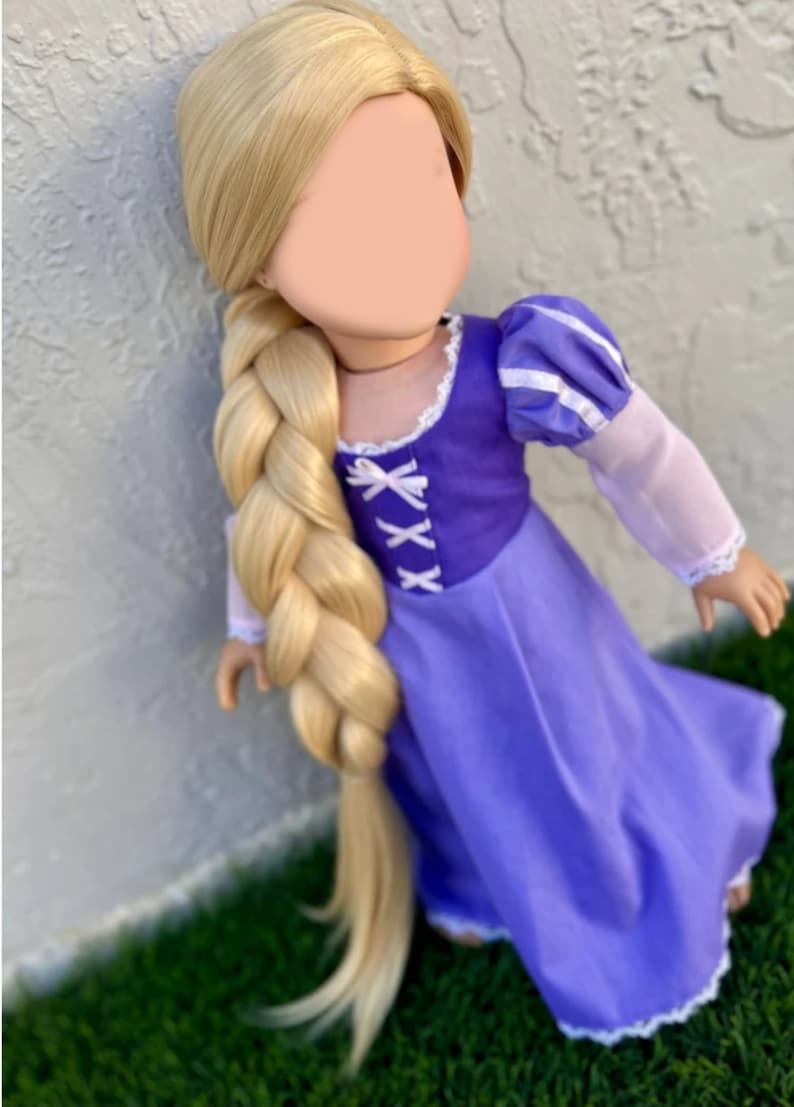 May include: A doll with long, blonde hair styled in a thick braid. The doll wears a purple dress with white accents and a bodice with criss-cross lacing. The sleeves are light pink with white trim. The doll is posed against a textured wall.