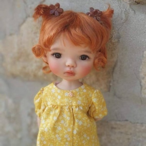 Puede incluir: Una muñeca pequeña con el pelo rojo peinado en coletas con horquillas marrones. La muñeca lleva un vestido amarillo con estampado floral blanco.