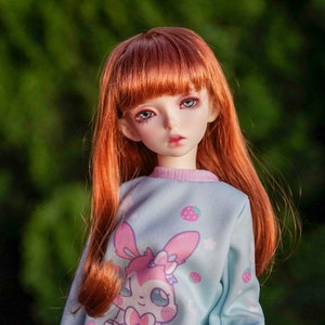 May include: A red-haired doll with blue eyes wearing a light blue sweatshirt with a pink bunny and strawberry print.