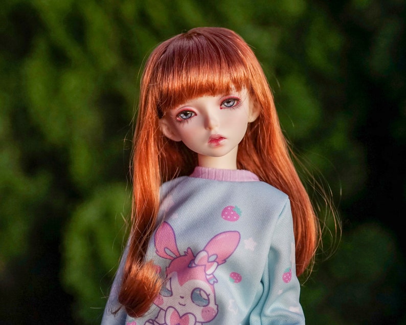 May include: A red-haired doll with blue eyes wearing a light blue sweatshirt with a pink bunny and strawberry print.