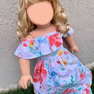 May include: A doll with blonde hair styled in an updo, wearing a white dress with a floral pattern. The dress features red, blue, and yellow flowers, with a ruffled neckline and sleeves. The doll is posed against a neutral background.