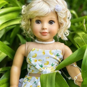 May include: A doll with blonde curly hair wearing a white and yellow lemon print dress and a white necklace. The doll is looking at the camera and has brown eyes.