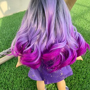 May include: A doll with long, wavy hair in shades of gray, lavender, and deep purple. The doll wears a purple dress with gold splatter details and blue sandals. The doll is standing on green grass.