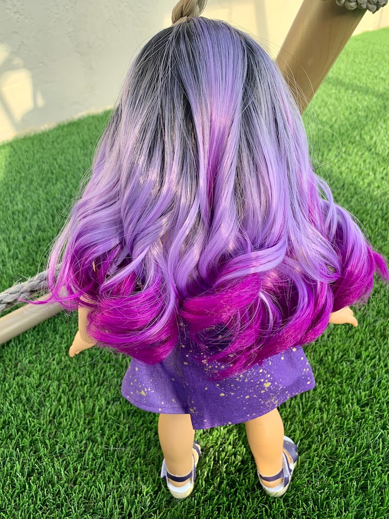 May include: A doll with long, wavy hair in shades of gray, lavender, and deep purple. The doll wears a purple dress with gold splatter details and blue sandals. The doll is standing on green grass.