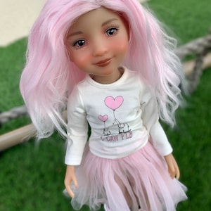 Puede incluir: Una muñeca con el pelo rosa que lleva una camisa blanca de manga larga con un corazón rosa y un gráfico de elefante. La camisa dice "Puedo volar". La muñeca también lleva una falda de tul rosa y zapatos rosas.