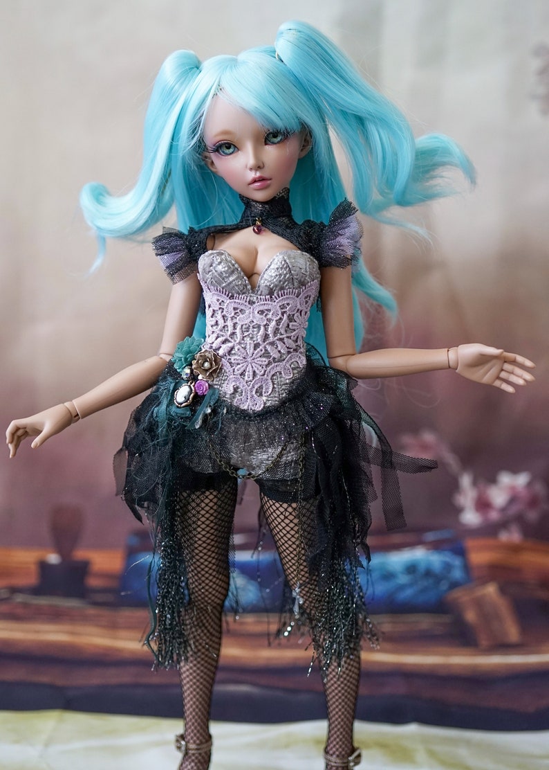 Puede incluir: Una mu&ntilde;eca con el pelo largo y azul brillante peinado en coletas, lleva un cors&eacute; negro y plateado con detalles de encaje, una falda de tul negro y medias de rejilla.
