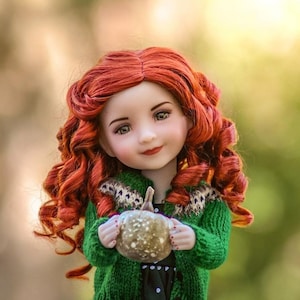 May include: A red-haired doll wearing a green sweater with a white and brown pattern holds a small, golden, pumpkin-shaped object.
