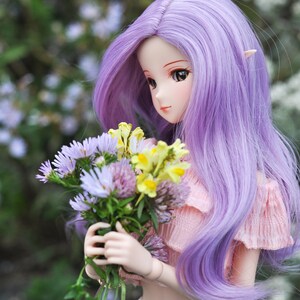 Op de afbeelding: Een pop met lang, paars haar houdt een boeket met paarse en gele bloemen vast. Ze draagt een roze top en een blauwe spijkerbroek.