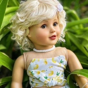 May include: A doll with blonde curly hair wearing a white and yellow lemon print top and skirt. The doll has blue eyes and is wearing a white necklace.