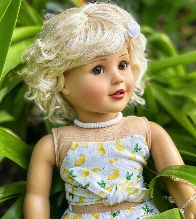 May include: A doll with blonde curly hair wearing a white and yellow lemon print top and skirt. The doll has blue eyes and is wearing a white necklace.
