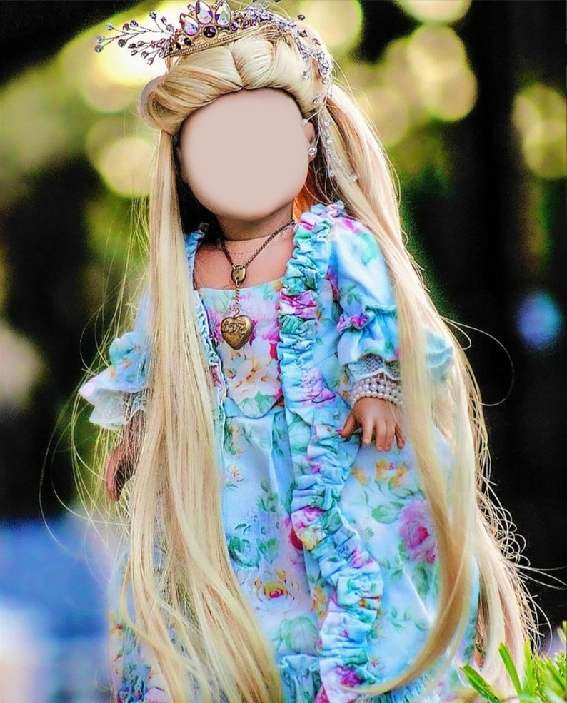 May include: A doll with long blonde hair, wearing a light blue floral dress with ruffled trim. The doll has a jeweled crown and a heart-shaped pendant necklace. The background is a blurred green.
