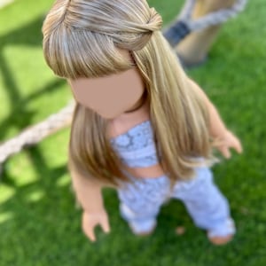 Puede incluir: Una mu&ntilde;eca con el pelo largo y rubio, peinado en una trenza, con un atuendo estampado en azul claro y blanco. La mu&ntilde;eca est&aacute; de pie sobre hierba verde.