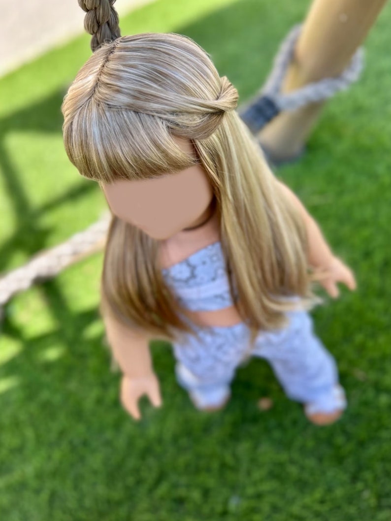 Puede incluir: Una mu&ntilde;eca con el pelo largo y rubio, peinado en una trenza, con un atuendo estampado en azul claro y blanco. La mu&ntilde;eca est&aacute; de pie sobre hierba verde.