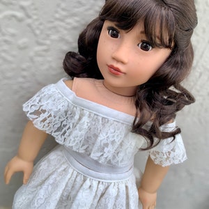 May include: A doll with long brown hair styled in curls and bangs, wearing a white off-the-shoulder lace dress.