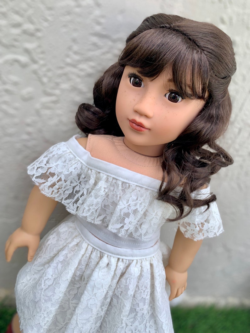 May include: A doll with long brown hair styled in curls and bangs, wearing a white off-the-shoulder lace dress.