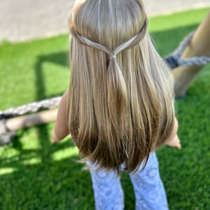 Puede incluir: Una mu&ntilde;eca con el pelo largo y rubio, peinado en una trenza con una peque&ntilde;a coleta en la parte posterior de la cabeza. La mu&ntilde;eca lleva pantalones blancos con un patr&oacute;n.