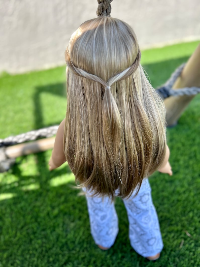 Puede incluir: Una mu&ntilde;eca con el pelo largo y rubio, peinado en una trenza con una peque&ntilde;a coleta en la parte posterior de la cabeza. La mu&ntilde;eca lleva pantalones blancos con un patr&oacute;n.
