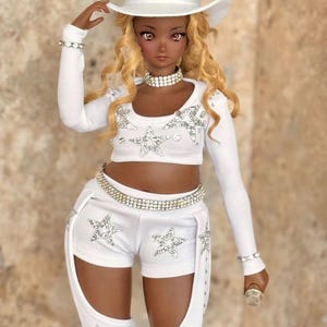 May include: A doll wearing a white cowboy hat, white long-sleeved shirt, white shorts with silver stars, and a silver belt. The doll has brown skin, long blonde hair, and is holding a microphone.