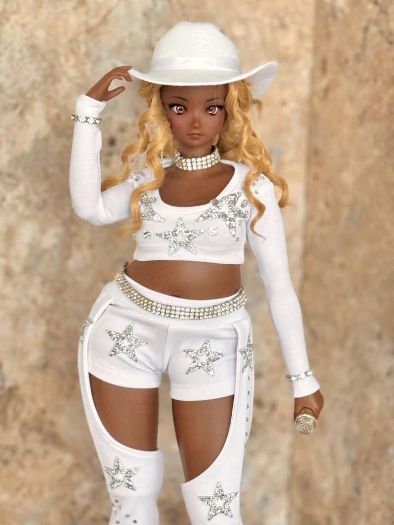 May include: A doll wearing a white cowboy hat, white long-sleeved shirt, white shorts with silver stars, and a silver belt. The doll has brown skin, long blonde hair, and is holding a microphone.