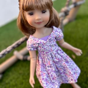 Puede incluir: Una muñeca con cabello castaño que lleva un vestido floral morado y blanco. La muñeca está de pie sobre hierba verde.