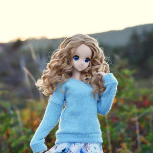 May include: A doll with long, curly blonde hair wearing a light blue sweater and a white skirt with a butterfly pattern.