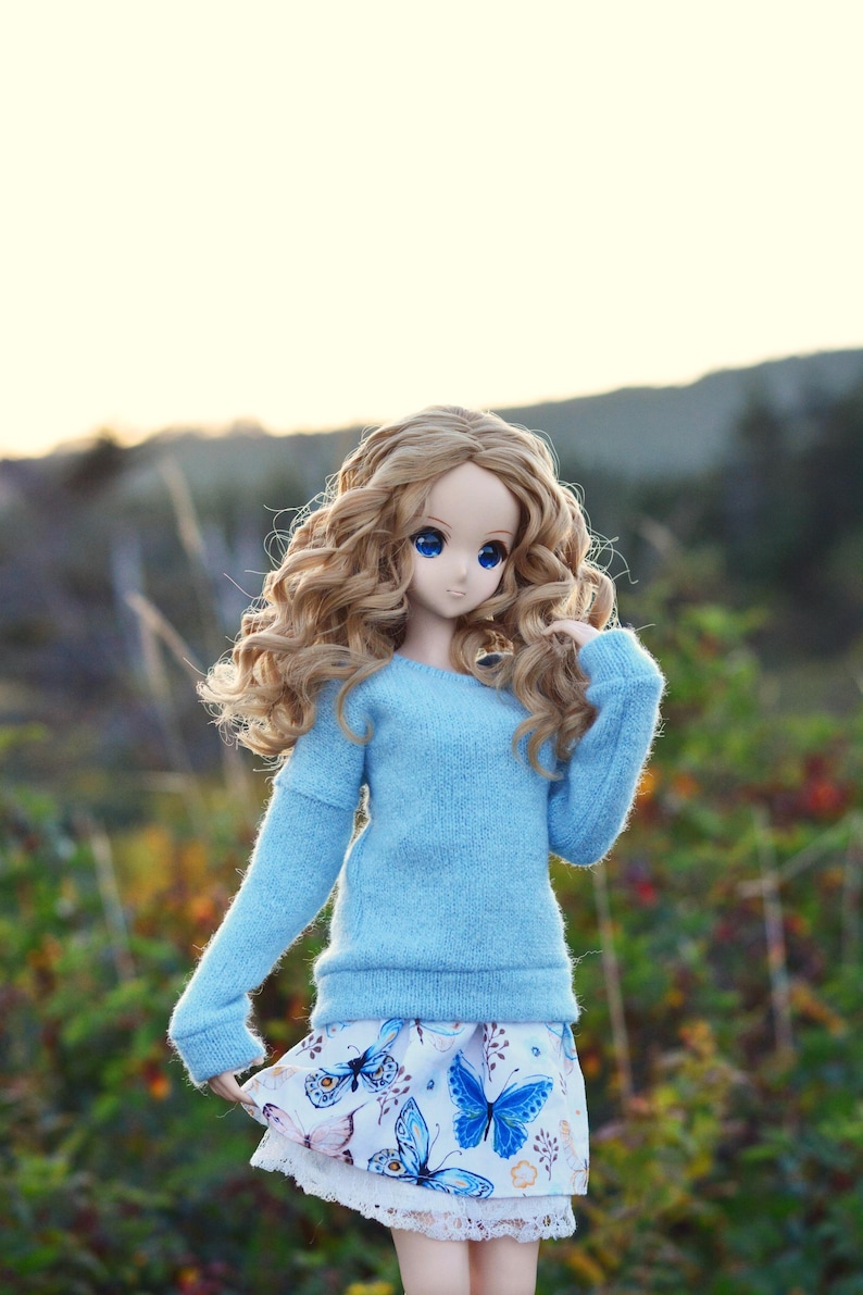 May include: A doll with long, curly blonde hair wearing a light blue sweater and a white skirt with a butterfly pattern.
