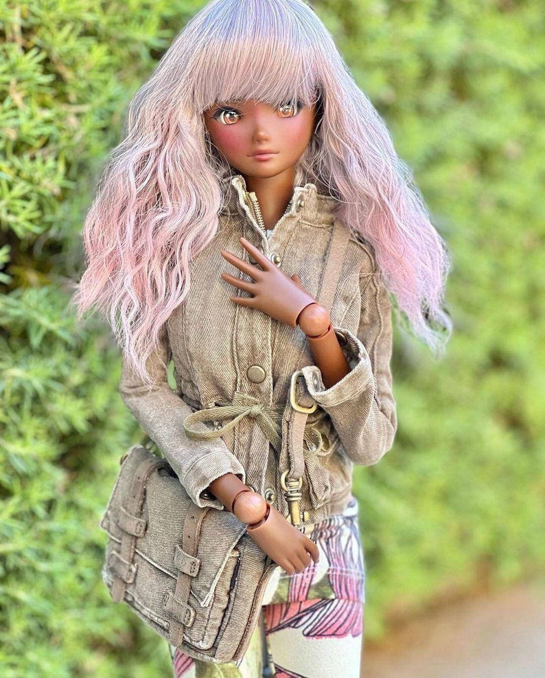 May include: A fashion doll with long, wavy, pink and lavender hair. The doll wears a tan jacket, patterned pants, and a matching satchel bag. The doll has a tan complexion and is posed in front of a green background.