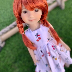 May include: A red-haired doll wearing a white dress with a cherry print. The doll has brown eyes and a sweet expression.