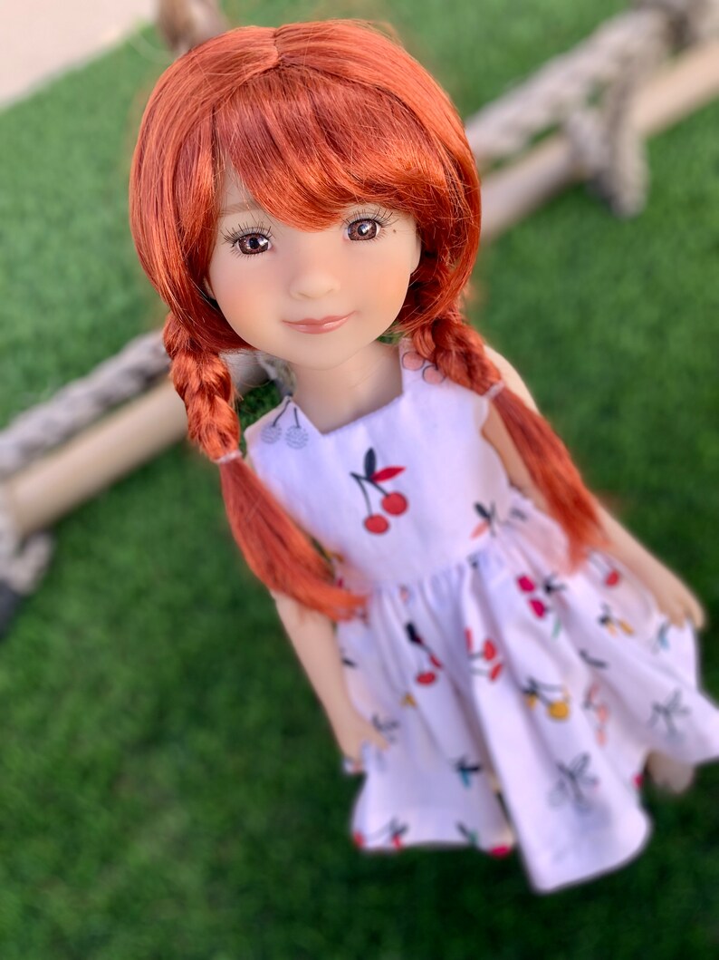 May include: A red-haired doll wearing a white dress with a cherry print. The doll has brown eyes and a sweet expression.
