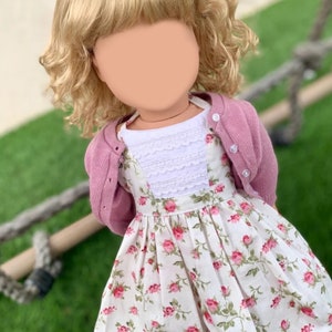 Puede incluir: Una muñeca con un cárdigan rosa, una blusa blanca con ribete de encaje y un vestido floral con rosas rosas. La muñeca tiene el pelo rubio rizado y lleva zapatos rosas.