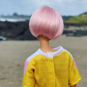 Peut inclure: Une poup&eacute;e avec des cheveux roses coiff&eacute;s en coupe au carr&eacute; portant une chemise jaune avec des accents blancs et roses.