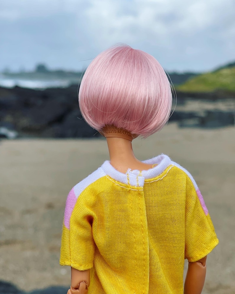 Peut inclure: Une poup&eacute;e avec des cheveux roses coiff&eacute;s en coupe au carr&eacute; portant une chemise jaune avec des accents blancs et roses.