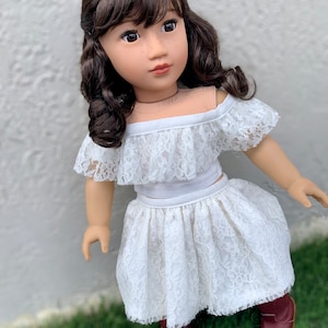 May include: A doll wearing a white lace off-the-shoulder top and skirt with brown boots. The doll has brown hair and brown eyes.