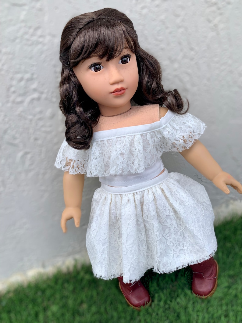 May include: A doll wearing a white lace off-the-shoulder top and skirt with brown boots. The doll has brown hair and brown eyes.