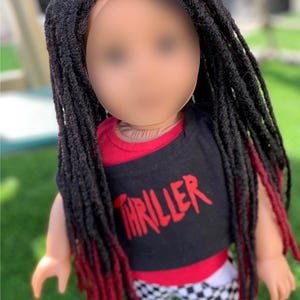 May include: A doll with long black dreadlocks with red tips. The doll is wearing a black t-shirt with the word "THRILLER" in red, a red and black top, and black and white checkered shorts.