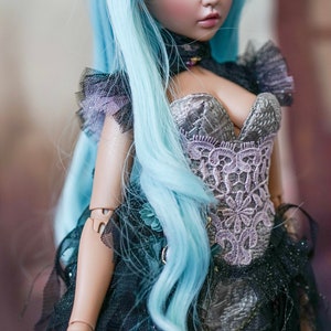 Puede incluir: Una mu&ntilde;eca con el pelo largo y azul claro que lleva un cors&eacute; de encaje negro y plateado con escote de coraz&oacute;n. La mu&ntilde;eca tiene una cara delicada con ojos azules y un ligero rubor rosado.