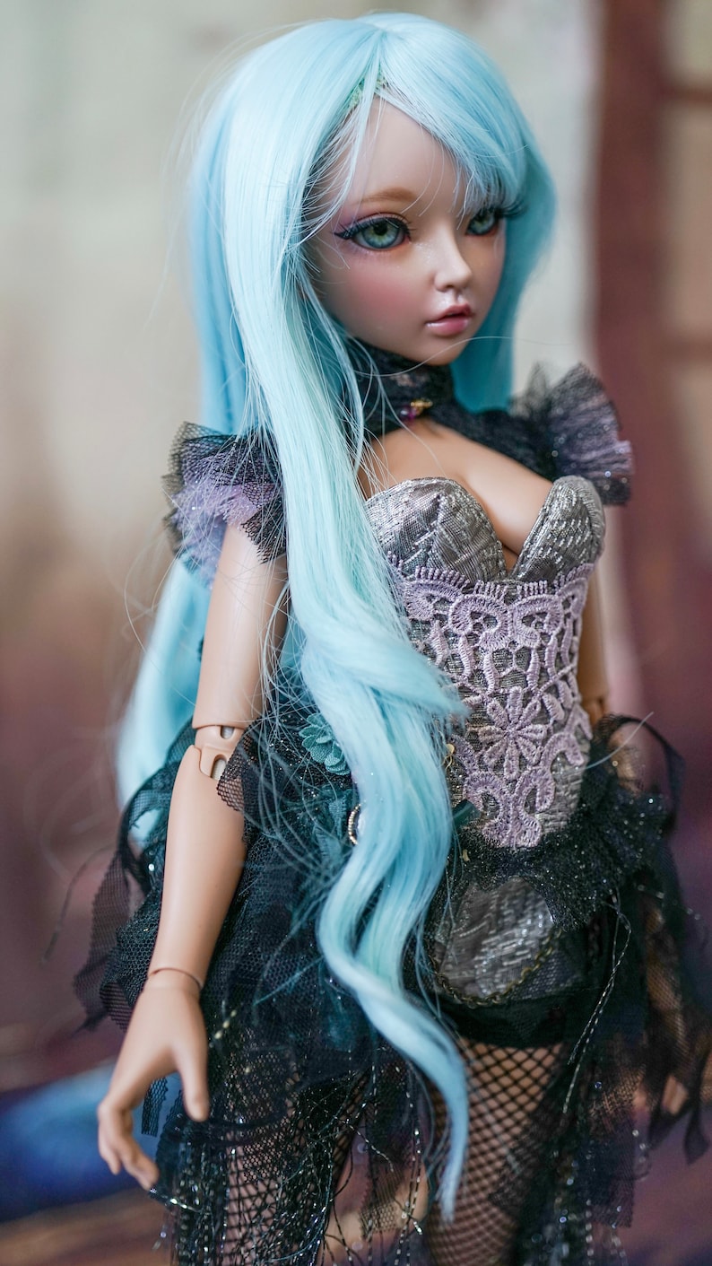Puede incluir: Una mu&ntilde;eca con el pelo largo y azul claro que lleva un cors&eacute; de encaje negro y plateado con escote de coraz&oacute;n. La mu&ntilde;eca tiene una cara delicada con ojos azules y un ligero rubor rosado.