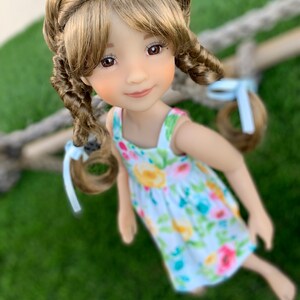 May include: A doll with long brown hair styled in braids with blue ribbons. The doll is wearing a white dress with a floral pattern and is looking at the camera.