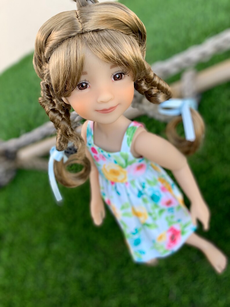 May include: A doll with long brown hair styled in braids with blue ribbons. The doll is wearing a white dress with a floral pattern and is looking at the camera.