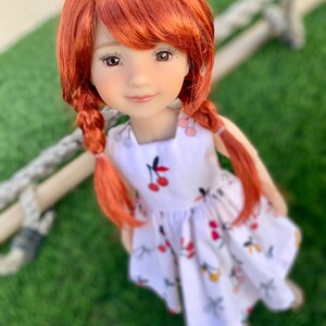 May include: A red-haired doll wearing a white dress with a cherry print. The doll has brown eyes and a sweet expression.