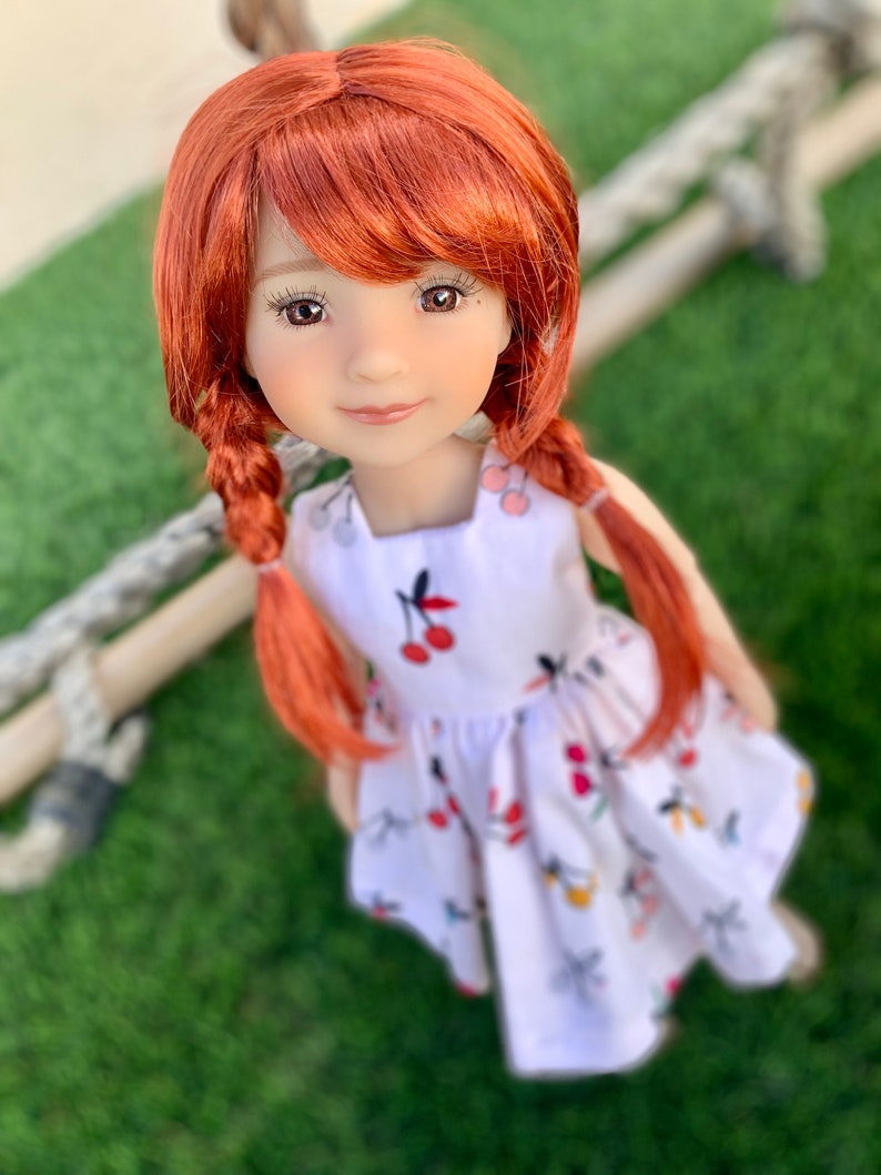 May include: A red-haired doll wearing a white dress with a cherry print. The doll has brown eyes and a sweet expression.