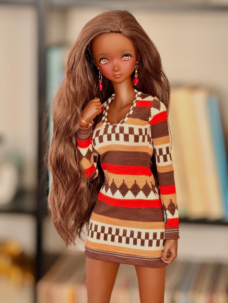 May include: A doll with long brown hair wearing a brown, red, and white patterned dress with long sleeves. The doll has brown skin and is wearing red earrings.