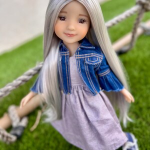 May include: A doll with long, silver hair and a gentle expression. She wears a blue denim jacket over a lavender dress and white shoes. The doll is posed against a green background, suggesting an outdoor setting.
