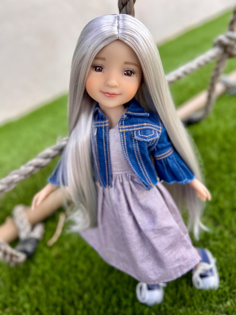 May include: A doll with long, silver hair and a gentle expression. She wears a blue denim jacket over a lavender dress and white shoes. The doll is posed against a green background, suggesting an outdoor setting.