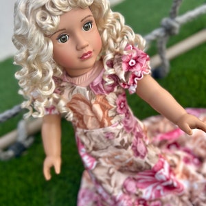May include: A doll with blonde curly hair wearing a floral dress with pink and brown flowers. The doll has green eyes and is wearing a light pink top.