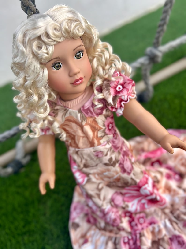 May include: A doll with blonde curly hair wearing a floral dress with pink and brown flowers. The doll has green eyes and is wearing a light pink top.