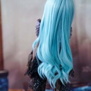 Puede incluir: Una mu&ntilde;eca con el pelo largo, ondulado y azul claro. La mu&ntilde;eca lleva un vestido negro con detalles brillantes.
