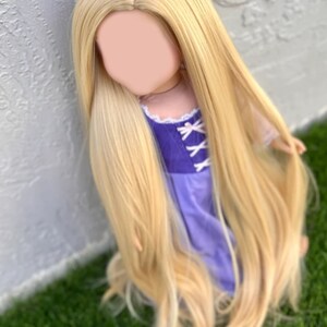 May include: A doll with long, flowing, blonde hair. The doll is wearing a purple dress with white accents. The hair is styled to cascade down the doll's body. The doll is positioned against a white wall and green grass.